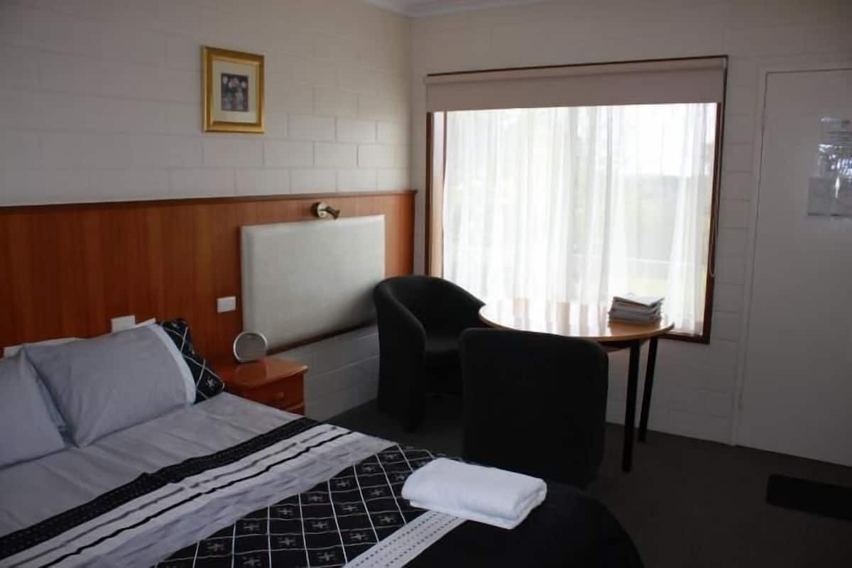 Habitación de hotel con cama, una silla, una mesita, ventana con cortinas y un cuadro en la pared.