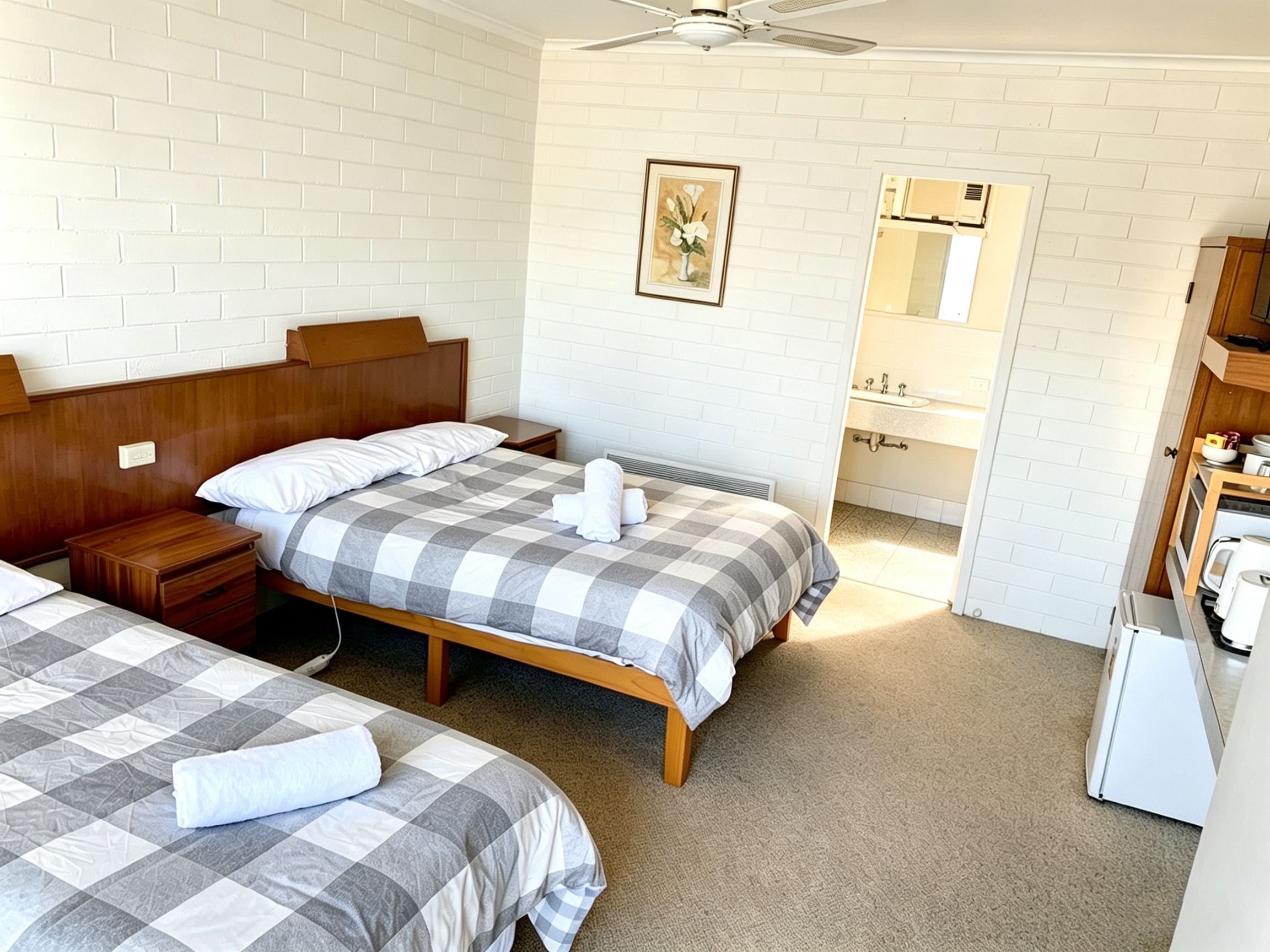 A hotel room with two beds, a wooden headboard, a bedside table, a nightstand, a ceiling fan, a bathroom with a sink and mirror, and a small kitchenette.