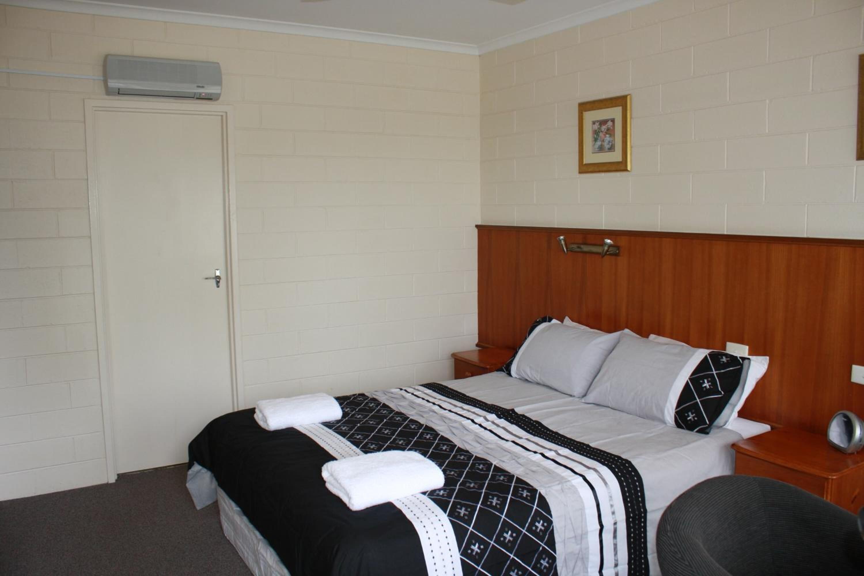 A hotel room with a bed, a wooden headboard, a bedside table, a chair, and a wall-mounted air conditioner.