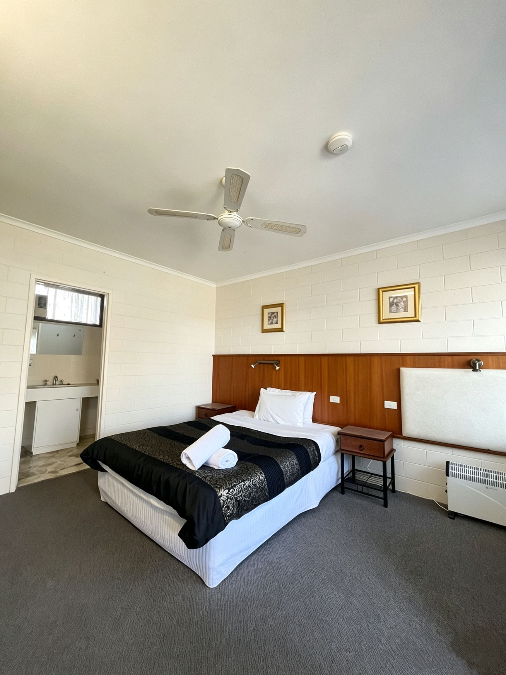 Een hotelkamer met een bed, nachtkastje, ventilator in het plafond, radiator en een klein raam.