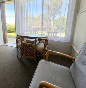 Una habitación con una mesa de vidrio y sillas de mimbre, un sofá gris y vistas a un jardín a través de una ventana con cortinas blancas.