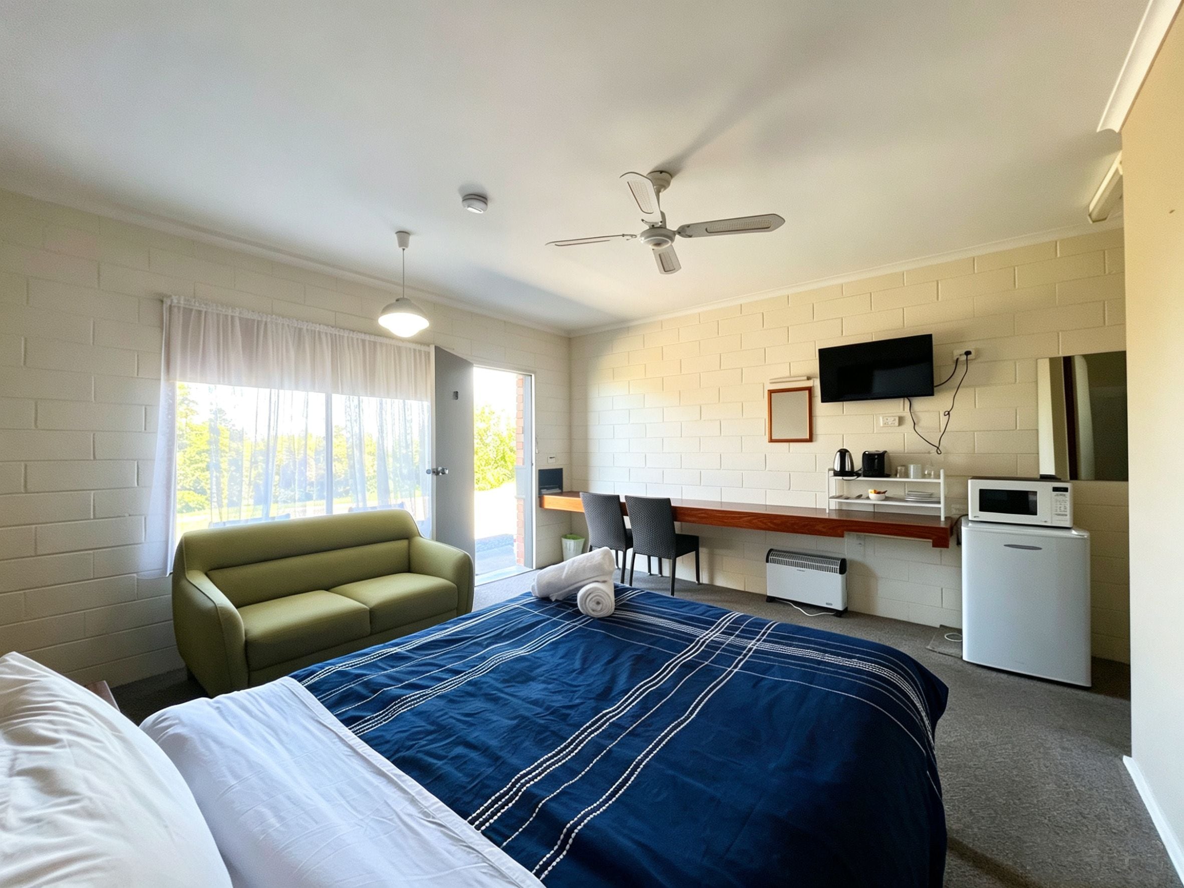 A hotel room with a bed, a sofa, a desk with a computer, a television, and a ceiling fan.