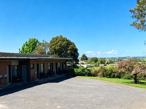 Un motel con un camino pavimentado, un área de estar al aire libre techada y vistas a un paisaje rural con árboles y campos.
