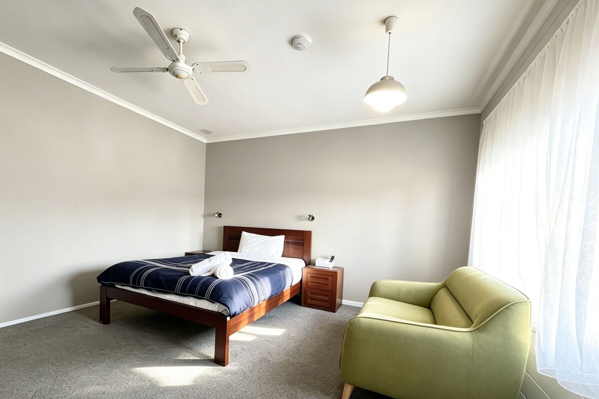 Un dormitorio con cama, mesita de noche, silla, ventilador de techo y ventana con cortinas.