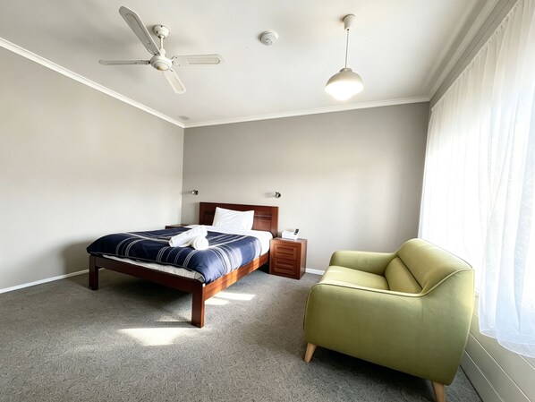 Un dormitorio con cama, mesita de noche, silla, ventilador de techo y ventana con cortinas.