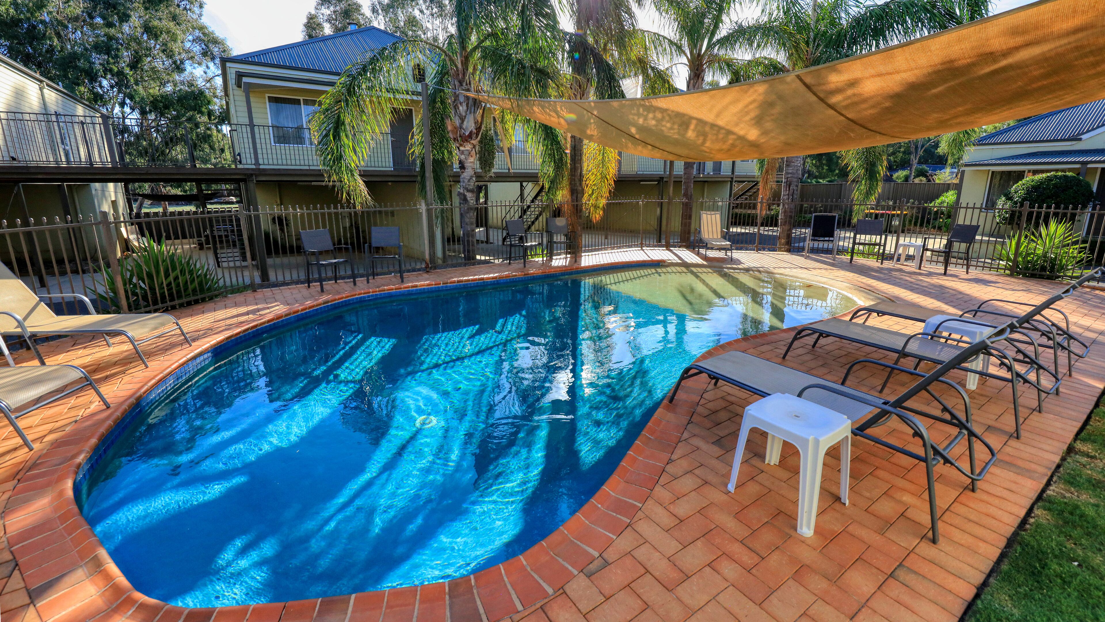 A swimming pool with a brick border, surrounded by lounge chairs and a shaded area.