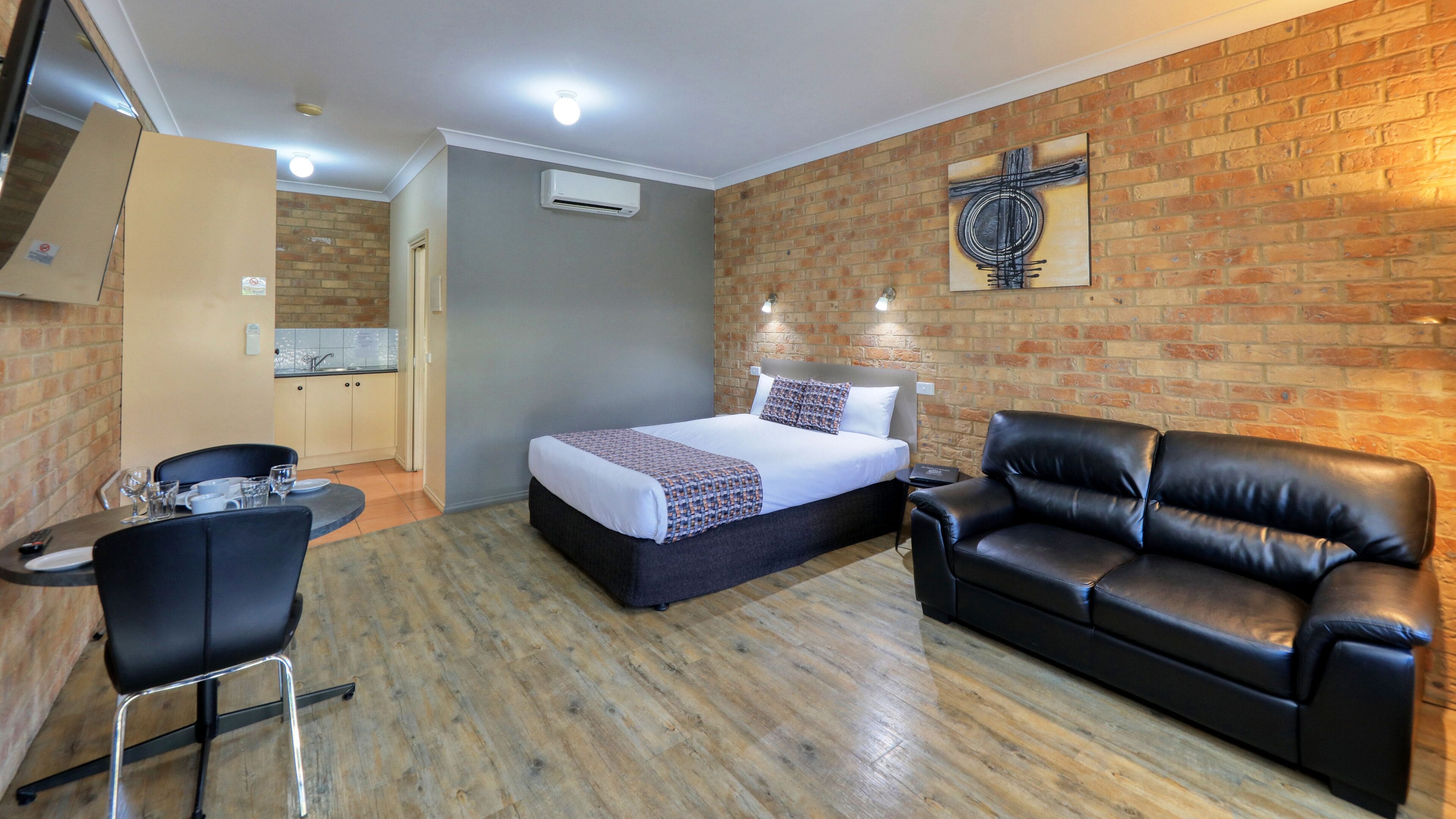 A compact hotel room with a bed, a leather sofa, a small table with a chair, and a kitchenette in the corner.