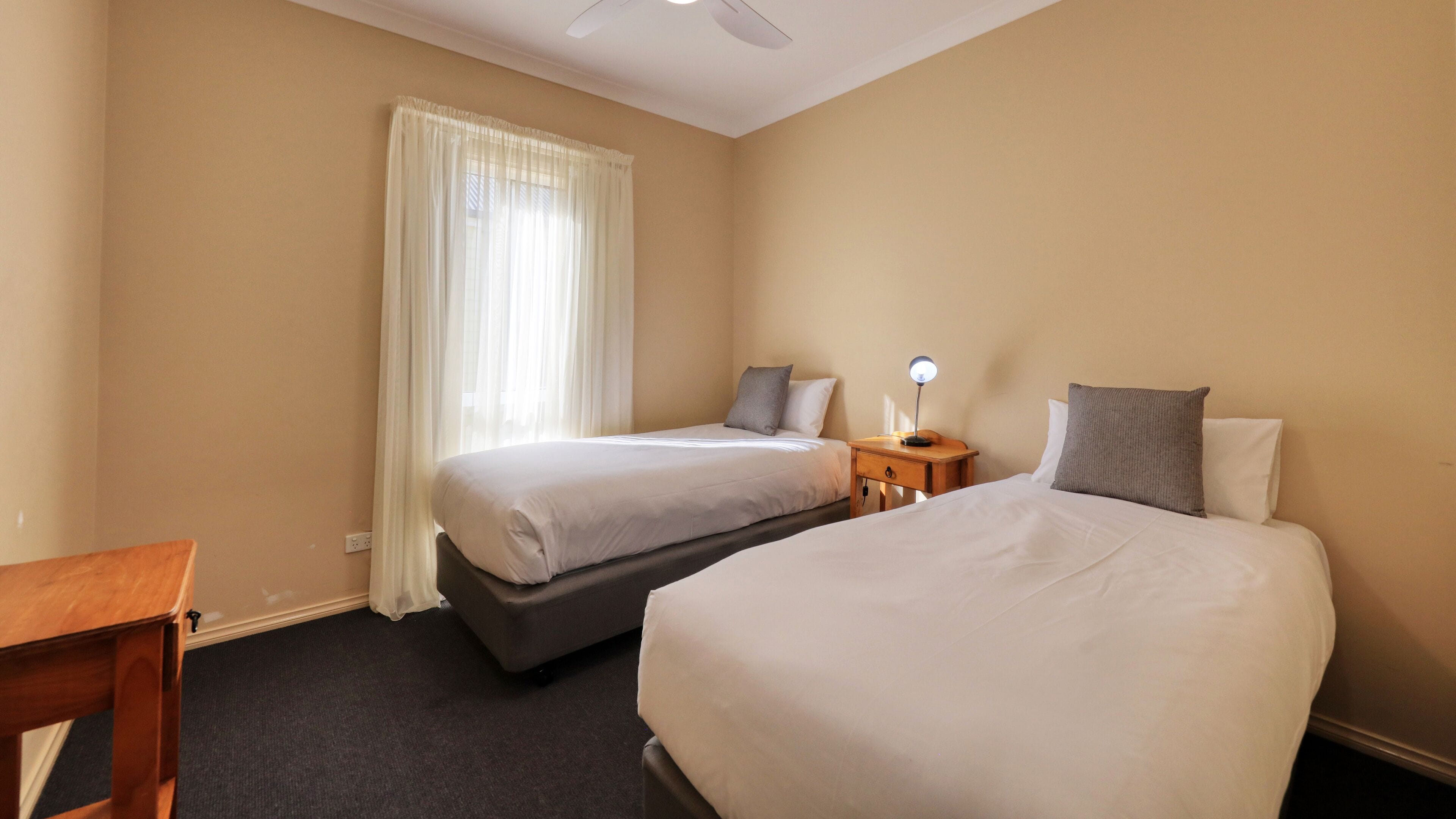 A hotel room with two beds, a desk, a nightstand with a lamp, and a window with curtains.