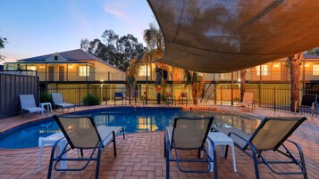 Una piscina con sillones de descanso, una zona cubierta y un edificio con varios balcones.