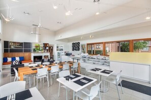Un espace repas moderne avec des tables et des chaises blanches, une cuisine en arrière-plan et un coin salon avec un canapé et une cheminée.