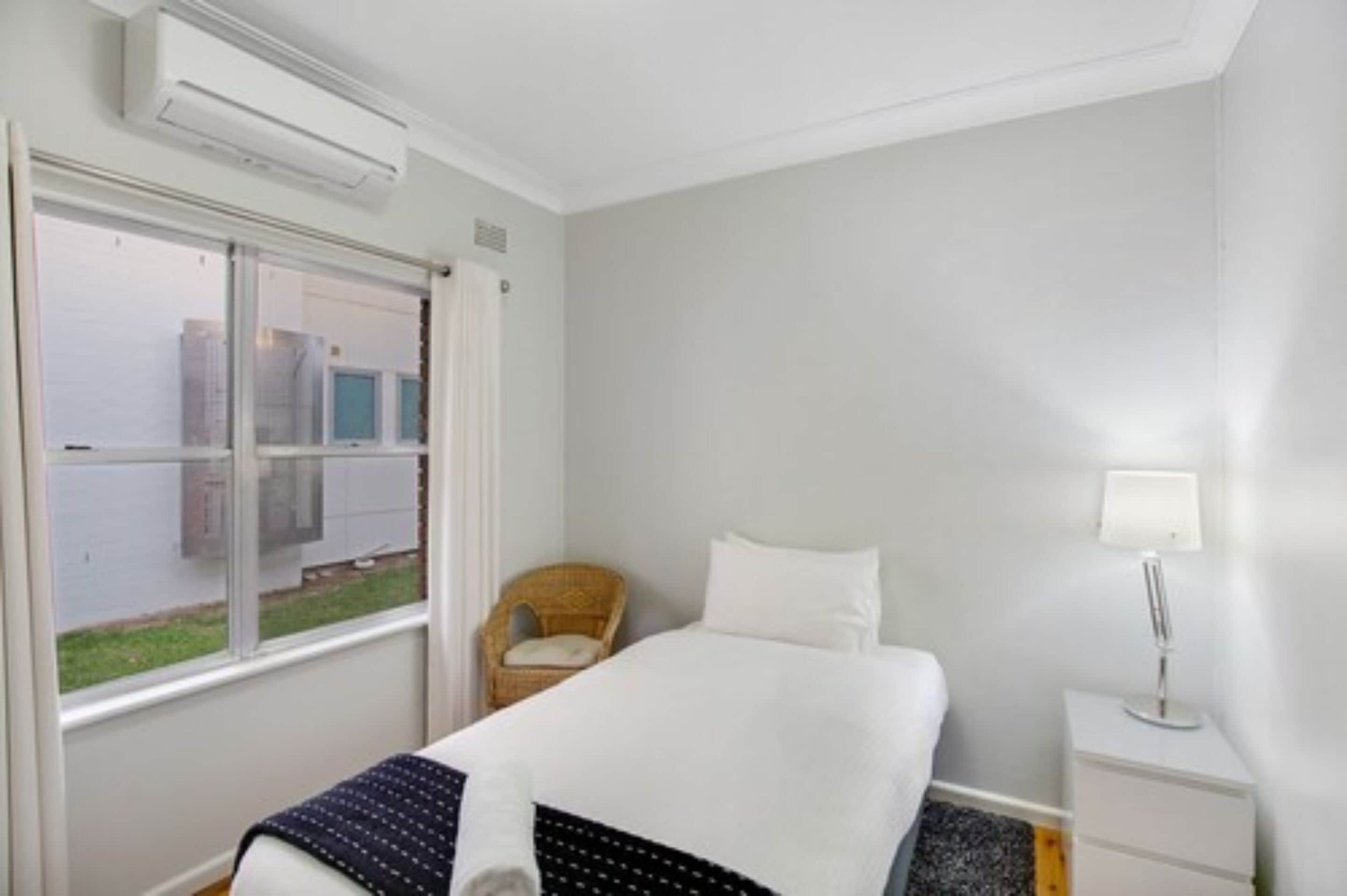 Hotellihuone, jossa on sänky, tuoli, yöpöytä lampun kanssa ja ikkuna, josta on näkymä rakennuksiin.