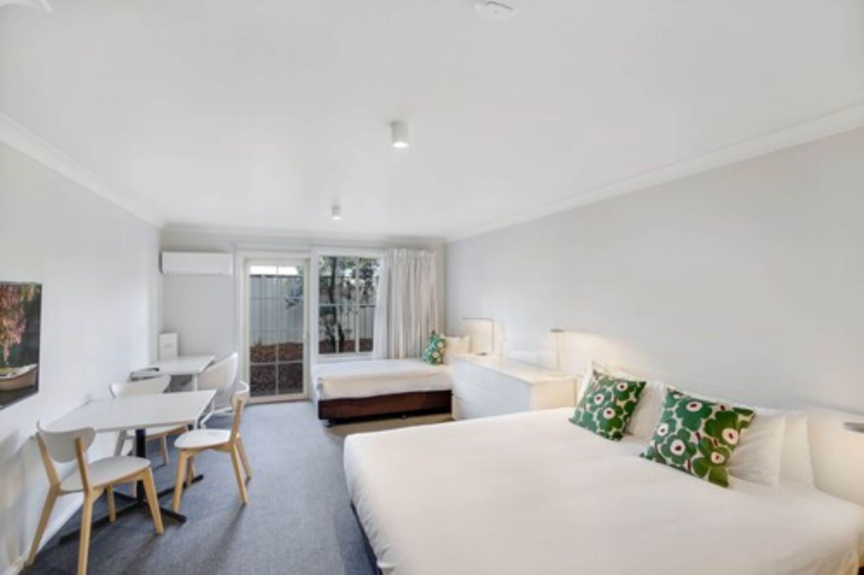 A hotel room with a bed, a desk, and a chair. There is a window with curtains and a view of trees outside.