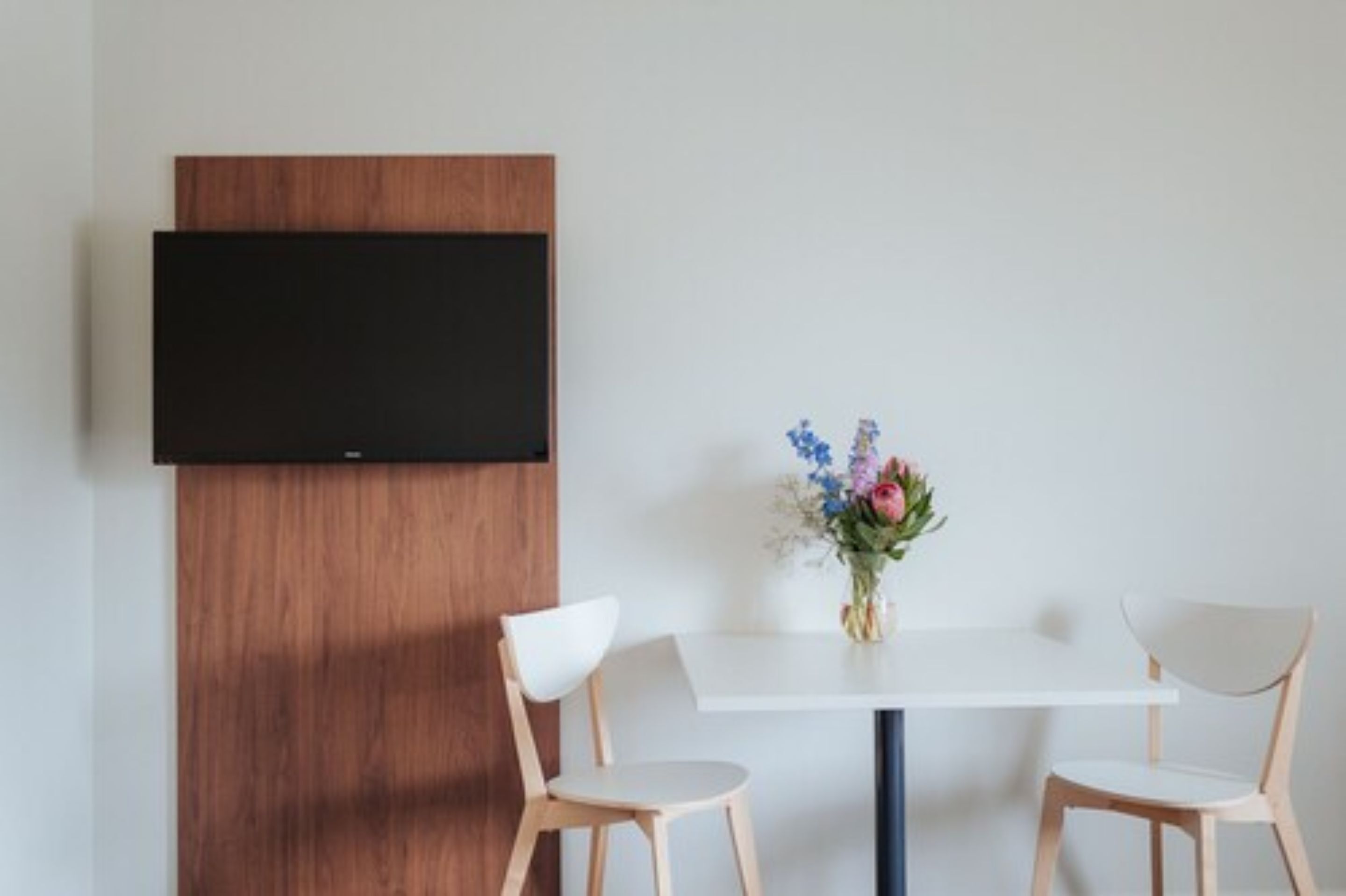 Een moderne kamer met een witte tafel, twee houten stoelen, een houten paneel en een flatscreen televisie aan de muur.