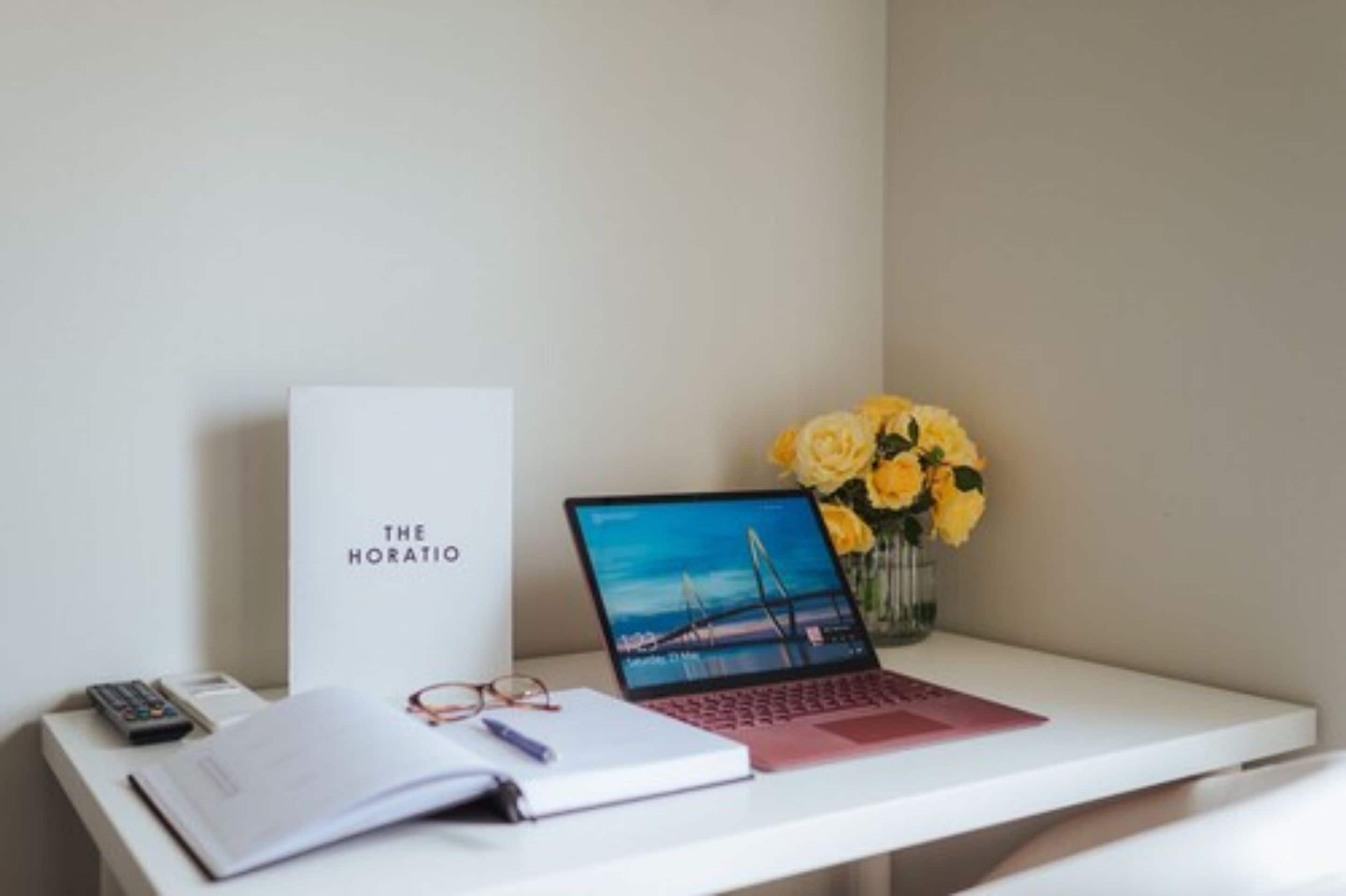 A desk with a laptop, a book, a pair of glasses, a calculator, and a vase with yellow flowers.