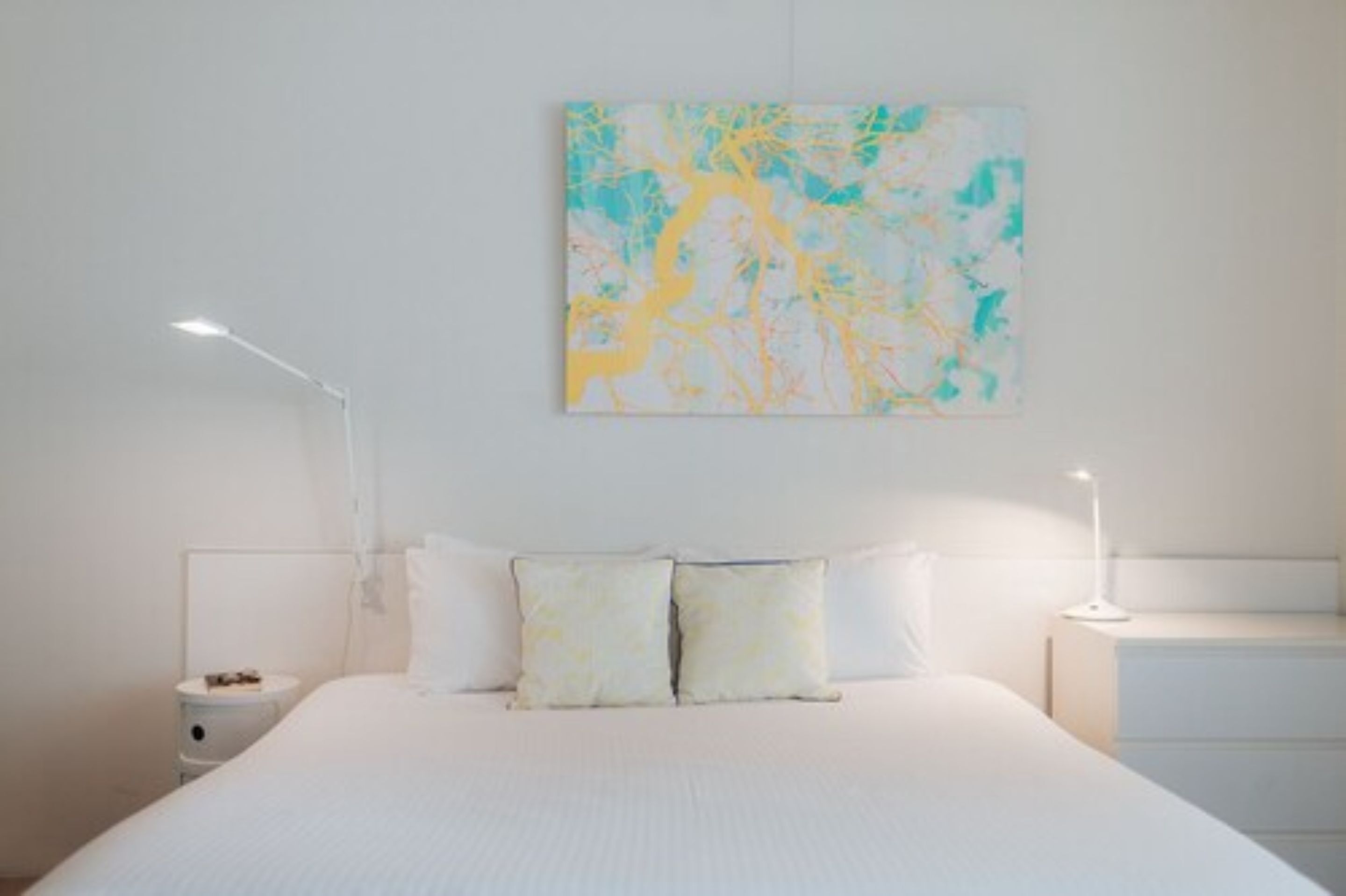 A bedroom with a large bed, two bedside tables, a wall-mounted abstract painting, and a white nightstand.