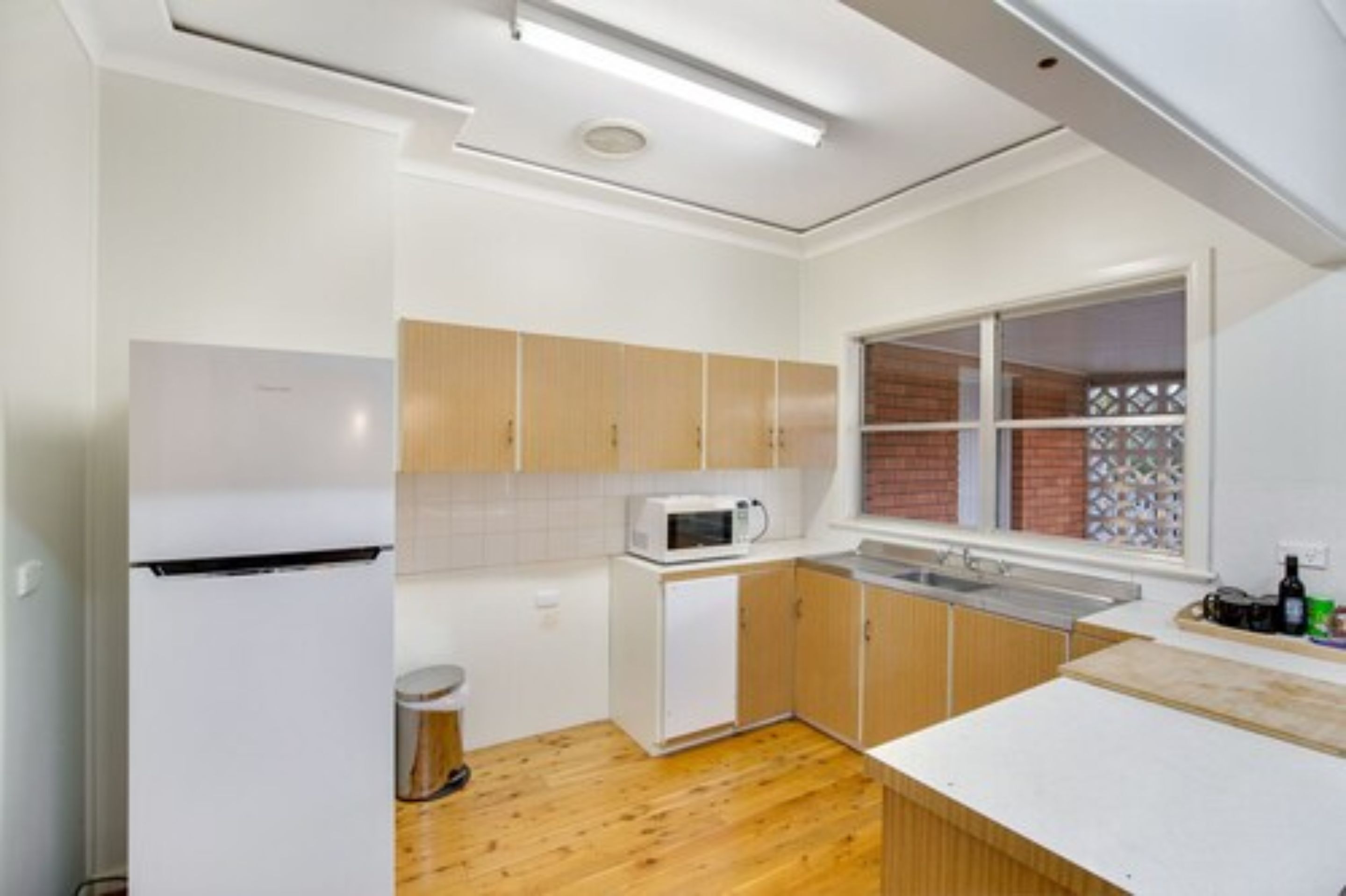 Cocina compacta con armarios blancos, un microondas y una ventana con vista a un muro de ladrillo.