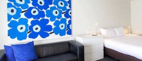 A hotel room with a black leather sofa, a bed with white bedding, a nightstand with a lamp, and a wall with a blue floral pattern.