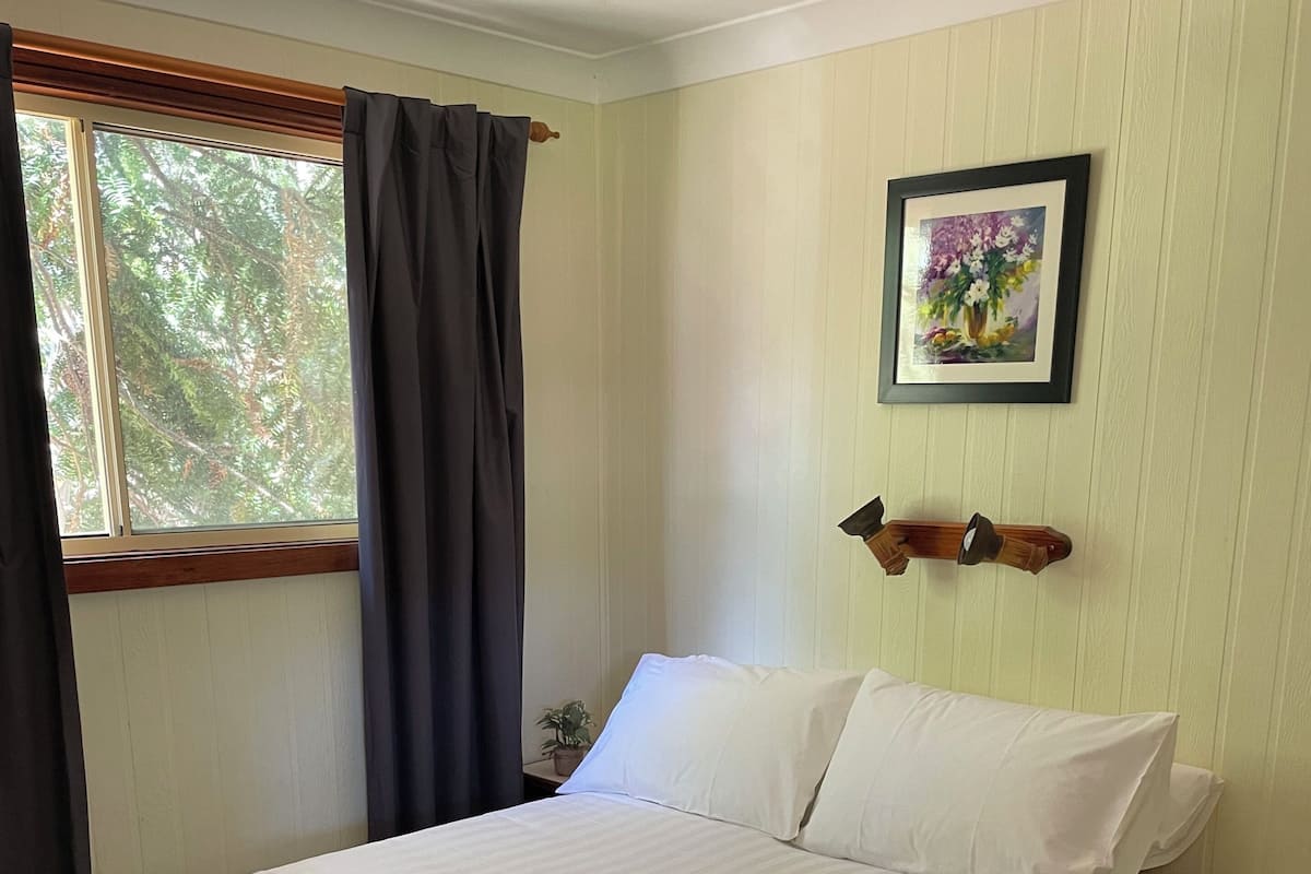 Una cama bien hecha con sábanas blancas y dos toallas dobladas, una ventana con cortinas, un cuadro colgado en la pared y una mesita de noche pequeña.
