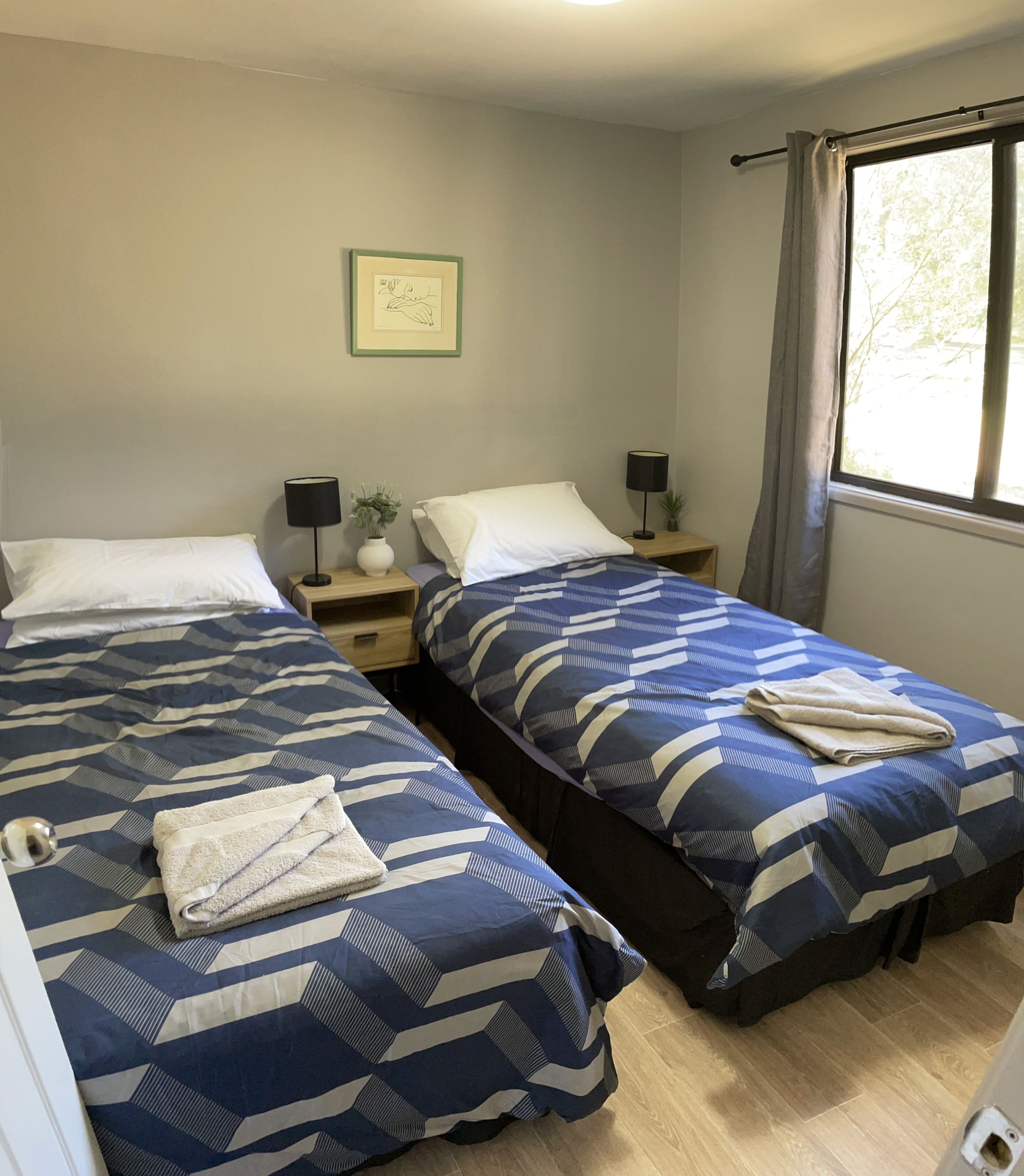 Une chambre double avec deux lits, des tables de chevet, des lampes, un tableau encadré et une fenêtre avec des rideaux.