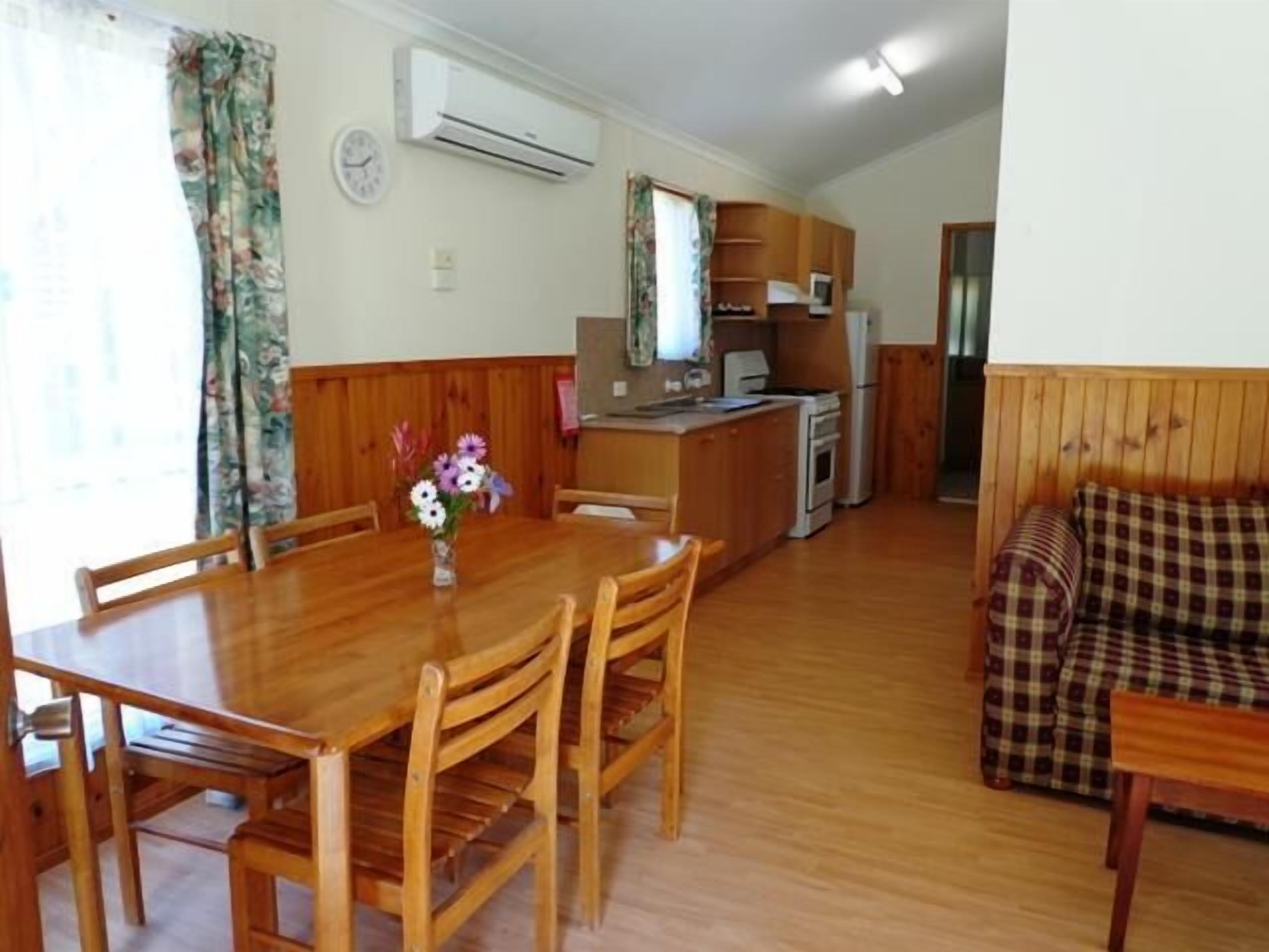 A dining area with a wooden table and chairs, a small kitchen area with a stove and refrigerator, and a living area with a sofa and a patterned cushion.
