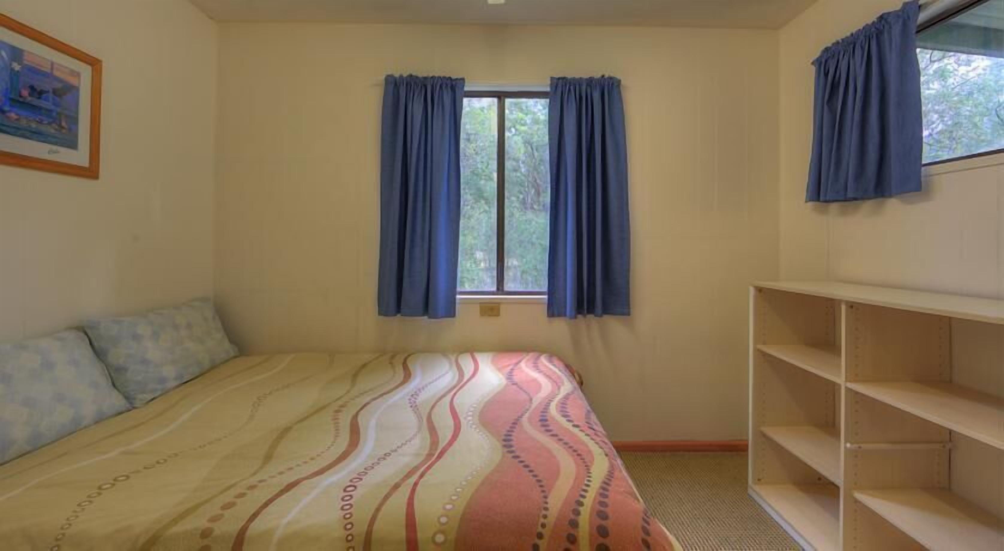A bedroom with a bed, a window with curtains, a shelf, and a framed picture on the wall.