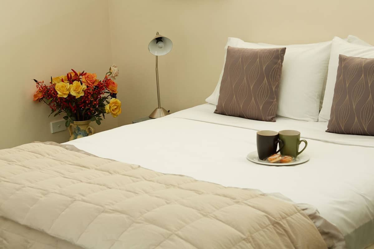 Cama bem arrumada com manta acolchoada, duas almofadas com padrão de folha, mesa de cabeceira com um vaso de flores e duas canecas sobre um prato.