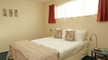 A neatly made bed with a beige comforter and pillows, a bedside table with a lamp and a book, and a window with red curtains.