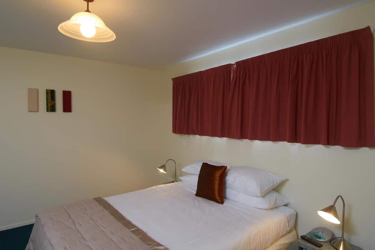 Cama bem arrumada com lençóis brancos e almofada marrom de destaque, cortina vermelha e luminária fixada na parede.