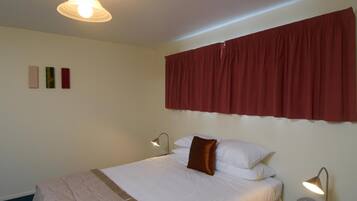 Una cama bien hecha con ropa de cama blanca y un cojín marrón decorativo, una cortina roja y una lámpara fijada a la pared.