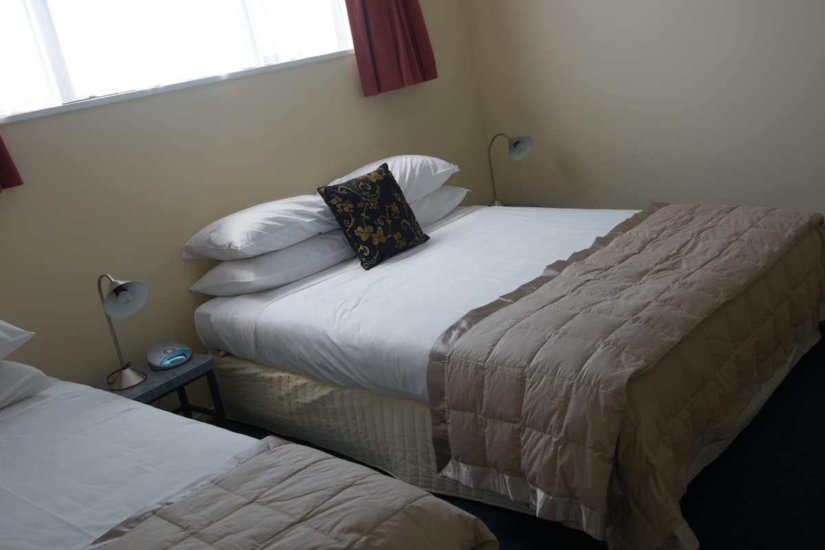 Quarto de hotel com duas camas, uma mesinha de cabeceira com abajur e uma janela com cortinas.