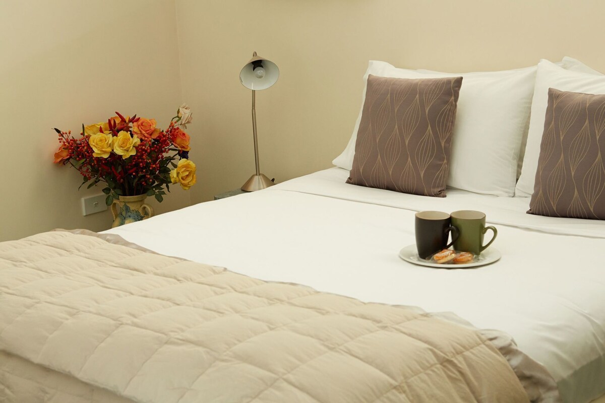 A neatly made bed with a quilted blanket, two pillows with a leaf pattern, a bedside table with a vase of flowers, and two mugs on a plate.