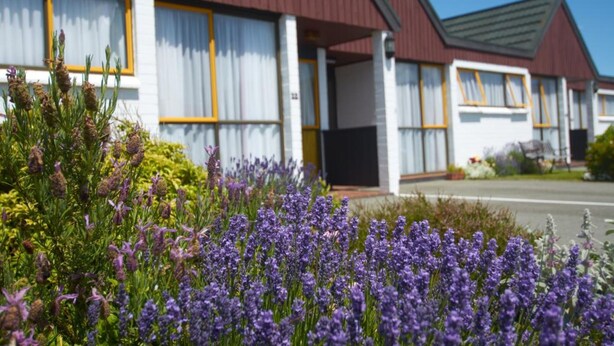 A row of motel buildings with white exteriors and red roofs, surrounded by a garden with purple flowers.