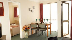 A hotel room with a wooden ceiling, a dining area with a table and chairs, a laptop on a side table, and a bed with a red curtain.