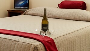 A hotel room with a bed, a laptop on a nightstand, a bottle of wine, and a red pillow.