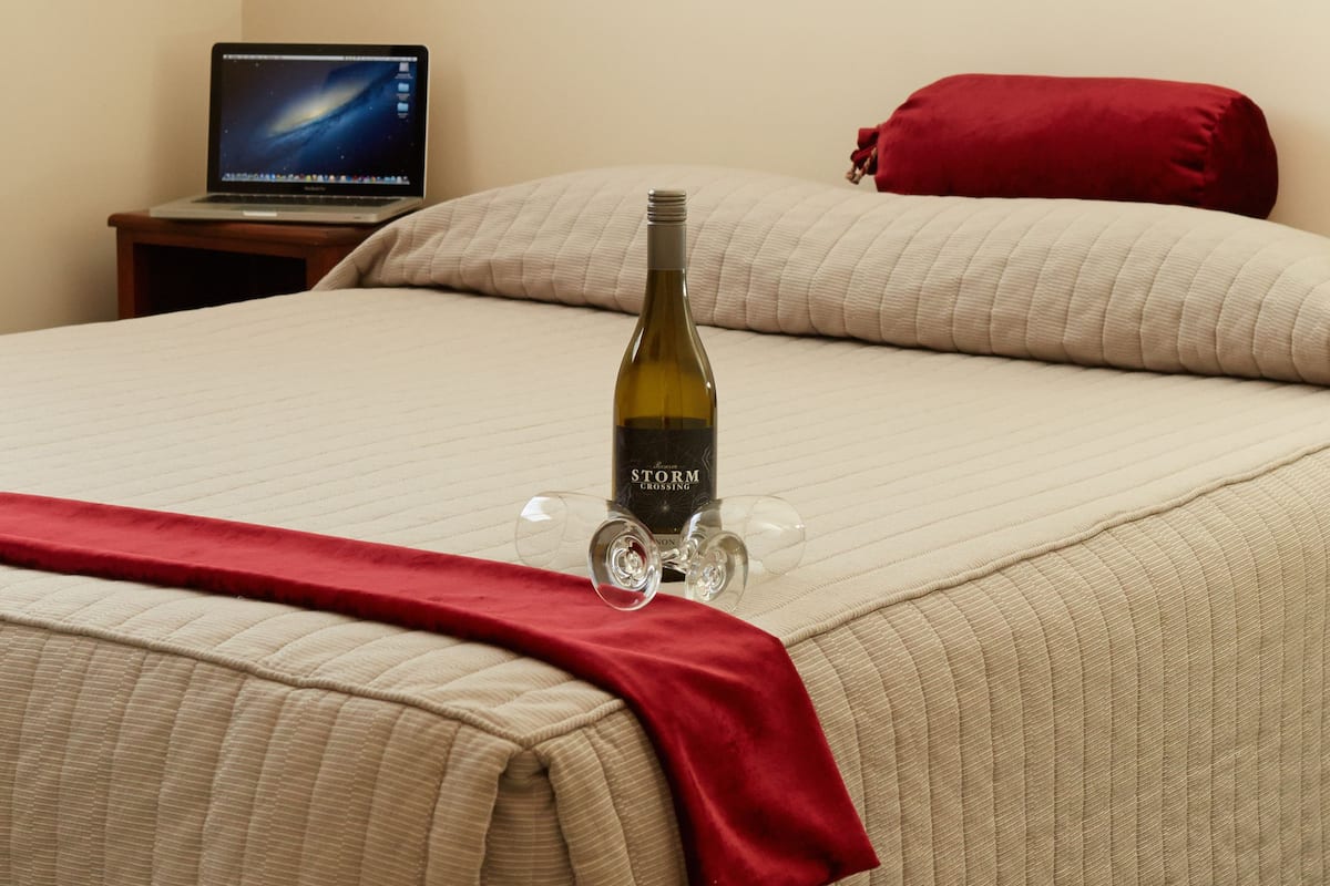 Quarto de hotel com uma cama, um laptop sobre uma mesinha de cabeceira, uma garrafa de vinho e uma almofada vermelha.