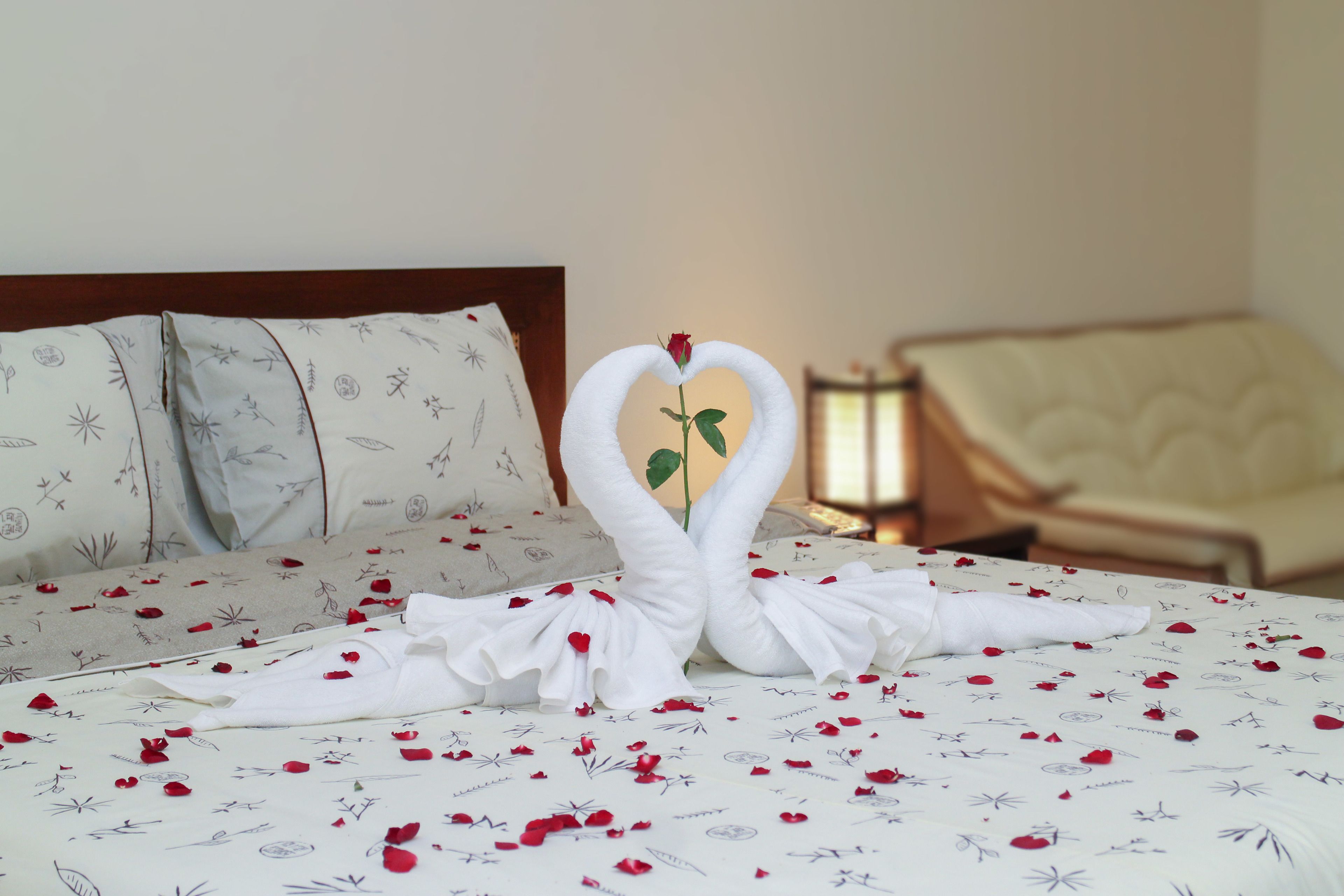 A bed with a heart-shaped towel arrangement and rose petals, a pillow with a floral pattern, and a chair in the background.
