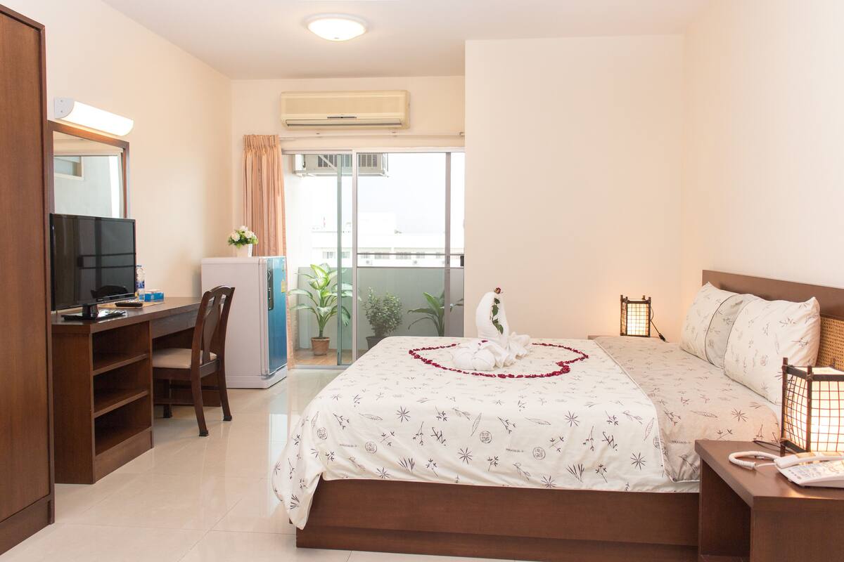 A hotel room with a bed, a desk with a computer, a chair, a TV, and a balcony with plants.
