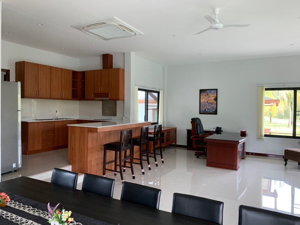 A modern kitchen with wooden cabinets, a central island with chairs, a dining table with chairs, a large window, and a ceiling fan.