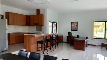 A modern kitchen with wooden cabinets, a central island with chairs, a dining table with chairs, a large window, and a ceiling fan.