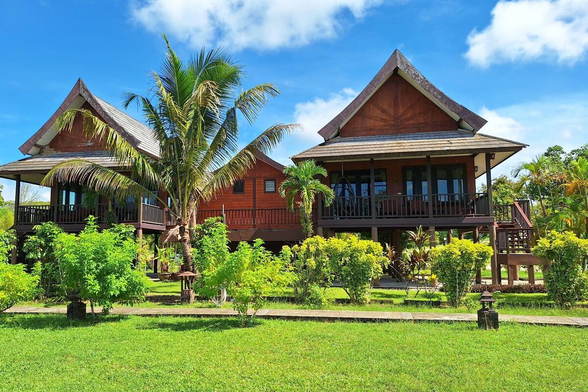 Een houten huis met twee verdiepingen en een balkon, omringd door weelderige begroeiing en palmbomen.