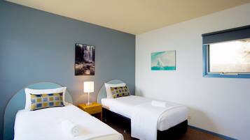 Quarto de hotel com duas camas de solteiro, mesa de cabeceira com luminária, pintura de uma cascata e janela com vista para um edifício.