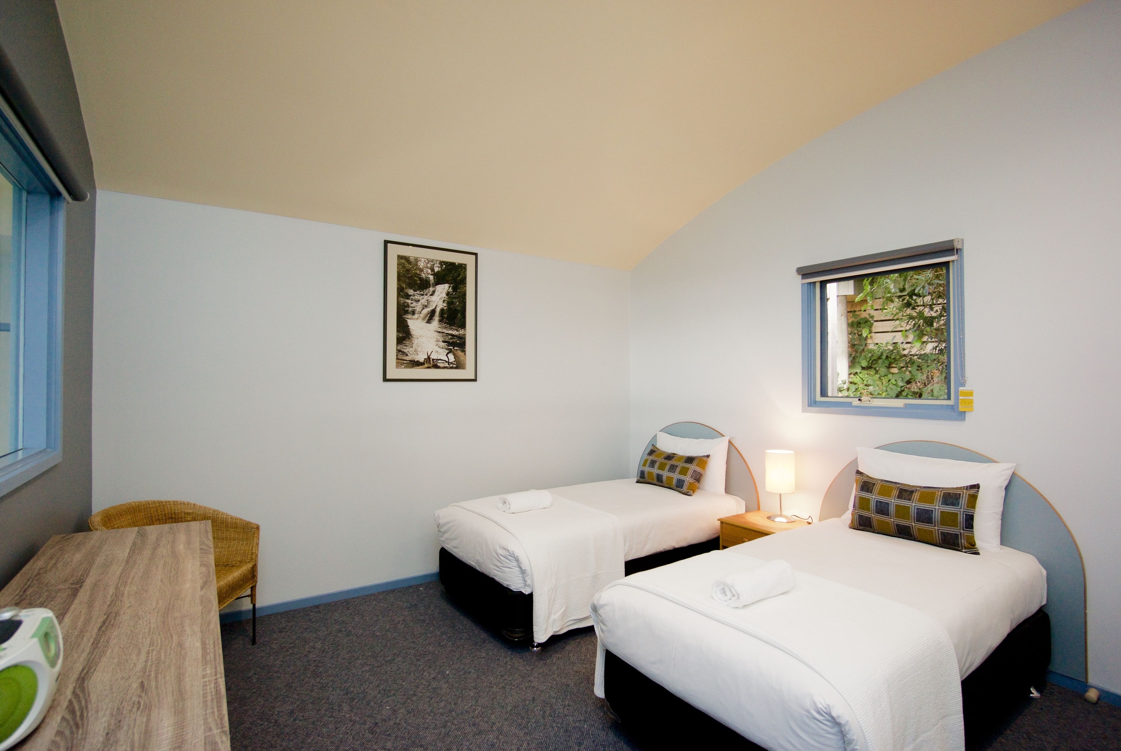 A room with two single beds, a wooden table, a chair, a framed picture on the wall, and a window with a view of greenery.