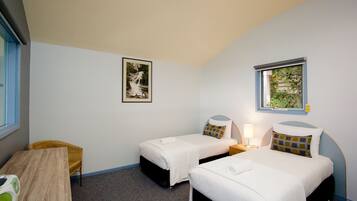 A room with two single beds, a wooden table, a chair, a framed picture on the wall, and a window with a view of greenery.