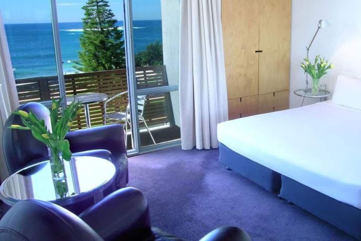Une chambre d’hôtel avec un grand lit, une petite table ornée d’un vase de fleurs, une chaise et une vue sur l’océan grâce à de grandes fenêtres.