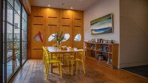 Un comedor con mesa de madera y sillas amarillas, una estantería con libros y objetos decorativos, y un ventanal con vistas al exterior.