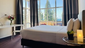 Habitación de hotel con una cama grande, un escritorio con silla, una mesita con una planta, una lámpara y vistas a árboles y al cielo a través de amplias ventanas.