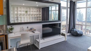 Una habitación moderna con un televisor grande, un escritorio blanco, estanterías con plantas y vistas al exterior.