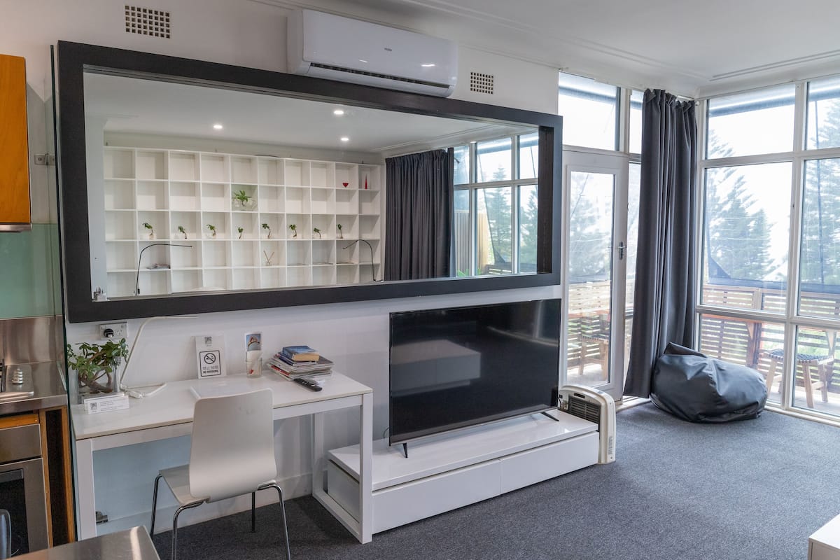 Une chambre moderne dotée d’une grande télévision, d’un bureau blanc, d’étagères ornées de plantes et offrant une vue sur l’extérieur.