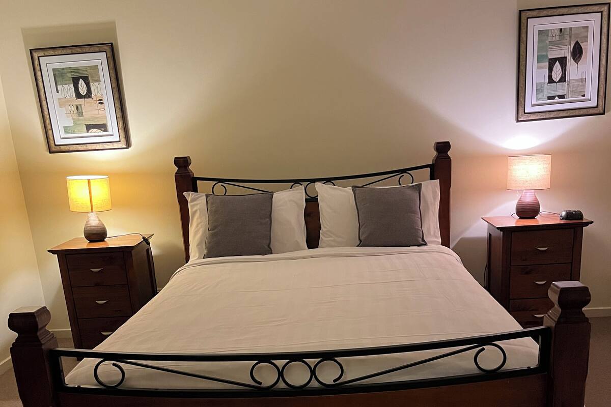 Een net opgemaakt bed met houten frame en hoofdbord, geflankeerd door twee nachtkastjes met lampen, in een kamer met effen muren en kunstwerken boven het bed.