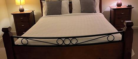 Una cama bien hecha con estructura y cabecera de madera, flanqueada por dos mesitas de noche con lámparas, en una habitación con paredes lisas y obras de arte enmarcadas sobre la cama.